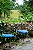 Delicate metal chairs with curved backrests and blue seat cushions in front of low, stone garden wall