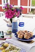 Bouquet in jug, beakers, plate of pastries and magazines on garden table