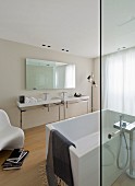 Free-standing white bathtub behind glass screen opposite twin washstands below large mirror in minimalist designer bathroom