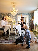 Child and dog playing and family sitting around festive dining table below lit chandelier in traditional dining room
