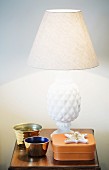 Gilt bowl and tin in front of table lamp with white fabric lampshade and pineapple-shaped base