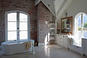 Freistehende Badewanne vor Fenstertür und weiße Einbauten im Badezimmer mit sichtbarem Ziegelmauerwerk