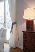 Table lamp hand-crafted from vintage coffee can on chest of drawers next to draped curtains on French windows