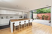 Bar stools with white loose covers in open-plan designer kitchen with open folding terrace doors to one side