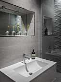 Badausschnitt - Waschbecken vor gefliester Wand, oberhalb Spiegel in Nische, davor Glasvasen mit Blumen auf Ablage