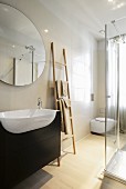 Trough-style sink with dark wood base cabinet, oval mirror on tiled wall, ladder used as towel rack and partially visible glass shower cabinet