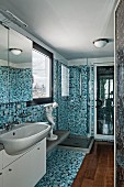 Narrow bathroom with mosaic tiles in various shades of blue, white washstand with mirrored cabinet and wooden floor section