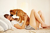Young woman lying on bed having face licked by pet dog