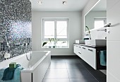 A shimmering, blue-and-white mosaic tiled wall behind a bathtub opposite a double washstand with a console basin and a wall-mounted, illuminated mirror