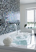 A shimmering mosaic tiled wall behind a bathtub with a bubble bath running