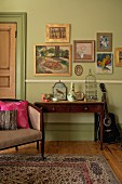 Small gallery of pictures on pastel green wall above ornaments and bird cage on writing desk; art nouveau armchair with various scatter cushions