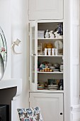 Jumble of crockery and china ornaments in fitted cupboard with open glass door