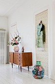 Vase of flowers on 50s, retro sideboard and demijohn full of scarves on floor below coat hooks on wallpapered door
