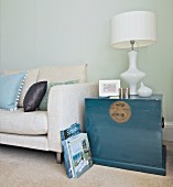 Elegant white table lamp on Oriental-style trunk painted blue-grey next to modern couch with pale linen cover against pastel wall