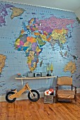 Antique armchair next to toy box and balance bike below bracket shelf on wall papered with map of world