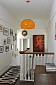 Orange pendant lamp above head of staircase on landing with white, wooden balustrade and gallery of pictures on wall