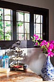 Delft vase of bougainvillea in front of Spanish, lattice windows, natural finds on cake stand on wooden surface