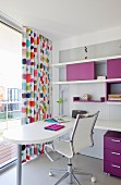 White desk and designer office chair in front of glass wall with multicoloured, polka-dot, floor-length curtains; shelving units with purple elements to one side