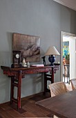 Table clock and table lamp on antique, carved, Oriental-style console table against grey-painted wall