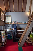 Young man in DIY loft-apartment kitchen with rustic ladder leading to gallery and retro fridge covered in colourful stickers on red carpet
