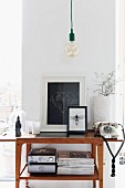 Minimalist, vintage pendant lamp above vintage telephone next to framed pictures on fifties-style console table against narrow section of wall