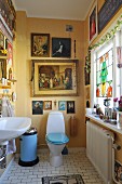 Yellow-painted bathroom with toilet, paintings with religious motifs on wall and ornaments on windowsill to one side