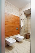 Bidet and toilet mounted on wood-clad projecting wall with glazed shower area in background