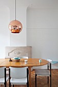 Pale wooden dining table and black and white chairs below pendant lamp with copper-coloured lampshade in traditional dining room with minimalist atmosphere