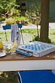 Altes Brettspiels und Erfrischungsgetränk auf Tisch im Garten; blaues Kissen und maritimes T-Shirt auf Gartenstühlen
