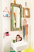 Jewellery hung from mirror frame with peeling paint and wire basket of bead necklaces and toiletries above small sink