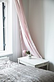 Shiny bedspread on bed, small table in corner and pink curtain