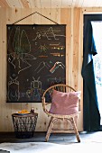 Rattanstuhl mit Kissen und Beistelltischchen vor aufgehängter Vintage-Lerntafel an naturbelassener Holzwand