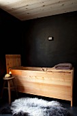 Wooden, trough-style bathtub against black wall, black and white sheepskin on black floor and lit candle