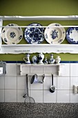 Kitchen utensils on small bracket shelf on tiled wall below carved shelf holding white and blue painted plates