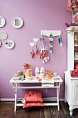 Weisser Wandtisch dekoriert, Kissen auf Fussgestell, vor fliederfarbener Wand, aufgehängte Dekowaren an Hakenleiste und bemalte Teller