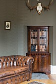 Antique, brown leather sofa, glass-fronted cabinet in niche below hunting trophy on wall with patterned green wallpaper