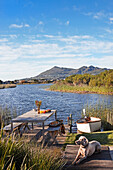 Rustikaler Sitzplatz mit Hund auf Holzdeck an idyllischem See mit Ruderboot am Ufer und Bergblick