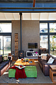 Pale green ottoman and leather sofa around coffee table, concrete partition with firewood stacked in niche and wood-burning stove in front of glass wall