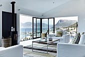 Elegant living room with white sofa set, delicate coffee table, partially glazed facade with sliding doors and panoramic view of mountainous coast