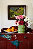 Vase of roses and dish of oranges on farmhouse cabinet with peeling paint on wall below landscape painting