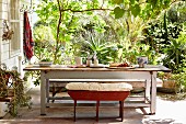 Bench with seat cushion at rustic table on terrace; tropical plants in sunny garden in background