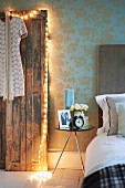 Dress hanging on removed vintage doors leaning against wall and decorated with fairy lights next to modern side table and partially visible bed
