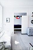 Sofa und Rollkoffer in Wohnraum mit Holzdielen, im Hintergrund Durchgang zum Schlafraum; Kunst an den Wänden