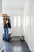 Woman in front of coat rack in rustic, white foyer with dark grey tiled floor and door mat with geometric pattern