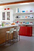 Kücheninsel und weiße Barhocker in offener Küche, an Wand gegenüber rote Schubladenkommode und weiße Regalböden mit buntem Geschirr