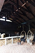 Two wicker hanging chairs with sheepskin blankets hanging from roof beam in barn with firewood stacked against wall