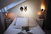 Bed below masks hung on wall flanked by lit table lamps on bedside tables in simple attic bedroom