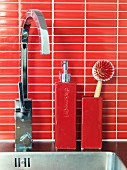 Ceramic soap dispenser and washing-up brush on stainless steel sink with angular, modern tap fittings and splashback of red mosaic tiles