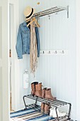 Offene Garderobe mit Ablage, auf Haken Jeansjacke und Strohhut, auf Metallbank Schuhe, vor Holzwand