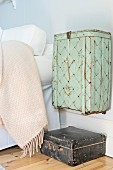 Old basket painted pastel green hung on wall above vintage, leather suitcase next to bed